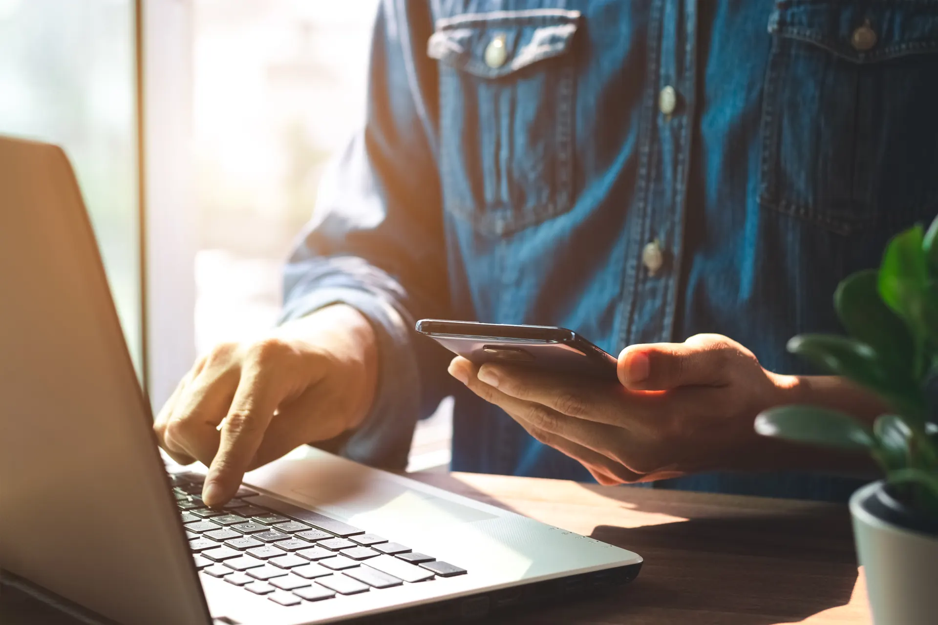 Person using a laptop and smartphone at a sunlit desk — contact UK Private Wealth Magazine