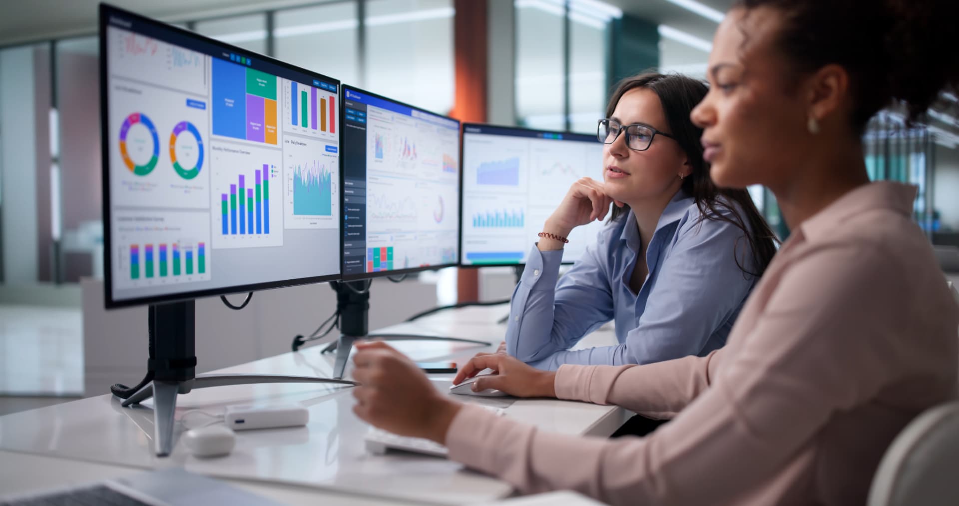 Two professionals reviewing data dashboards across multiple computer screens.
