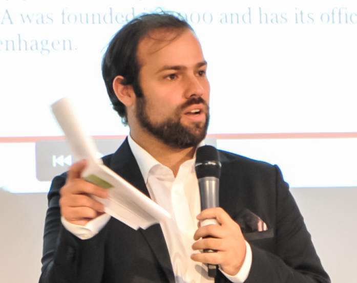 Alex Felman speaking at an event with a microphone.