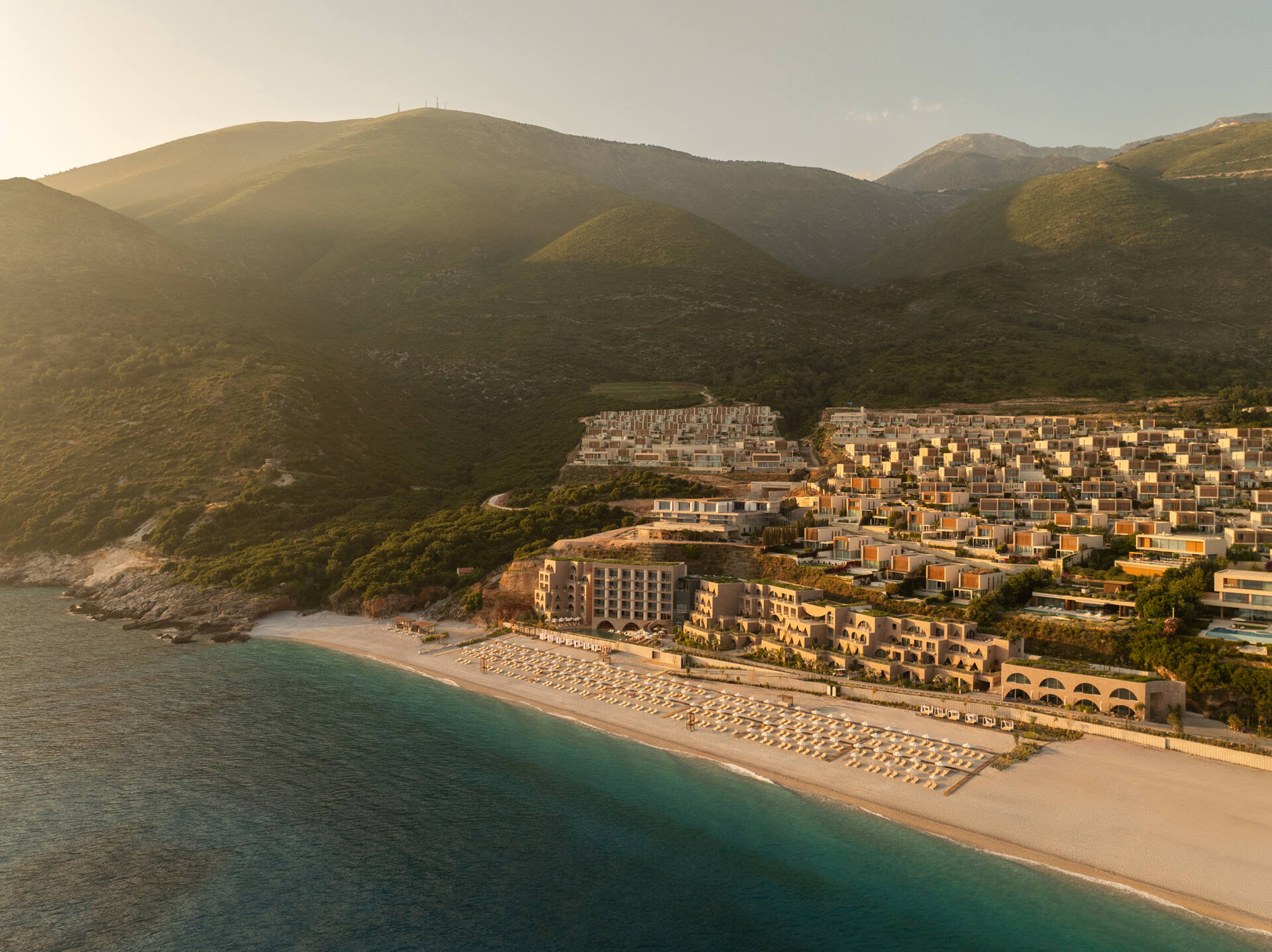 Aerial view at sunset of the Green Coast development on the Albanian Riviera, with terraced residences above a beach and turquoise sea.