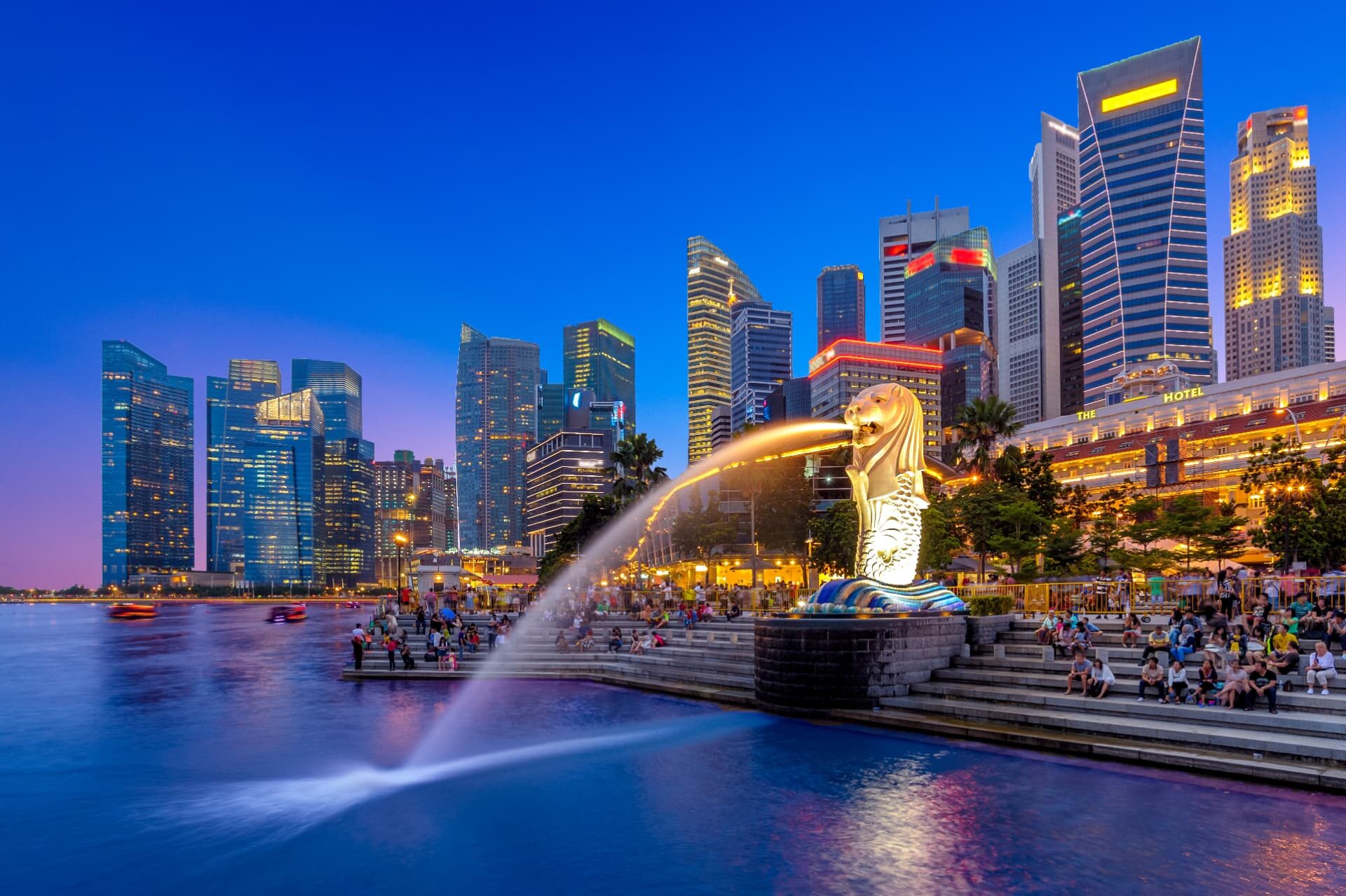 The Merlion fountain on the Marina Bay waterfront at dusk, with the lit towers of Singapore's financial district behind it.