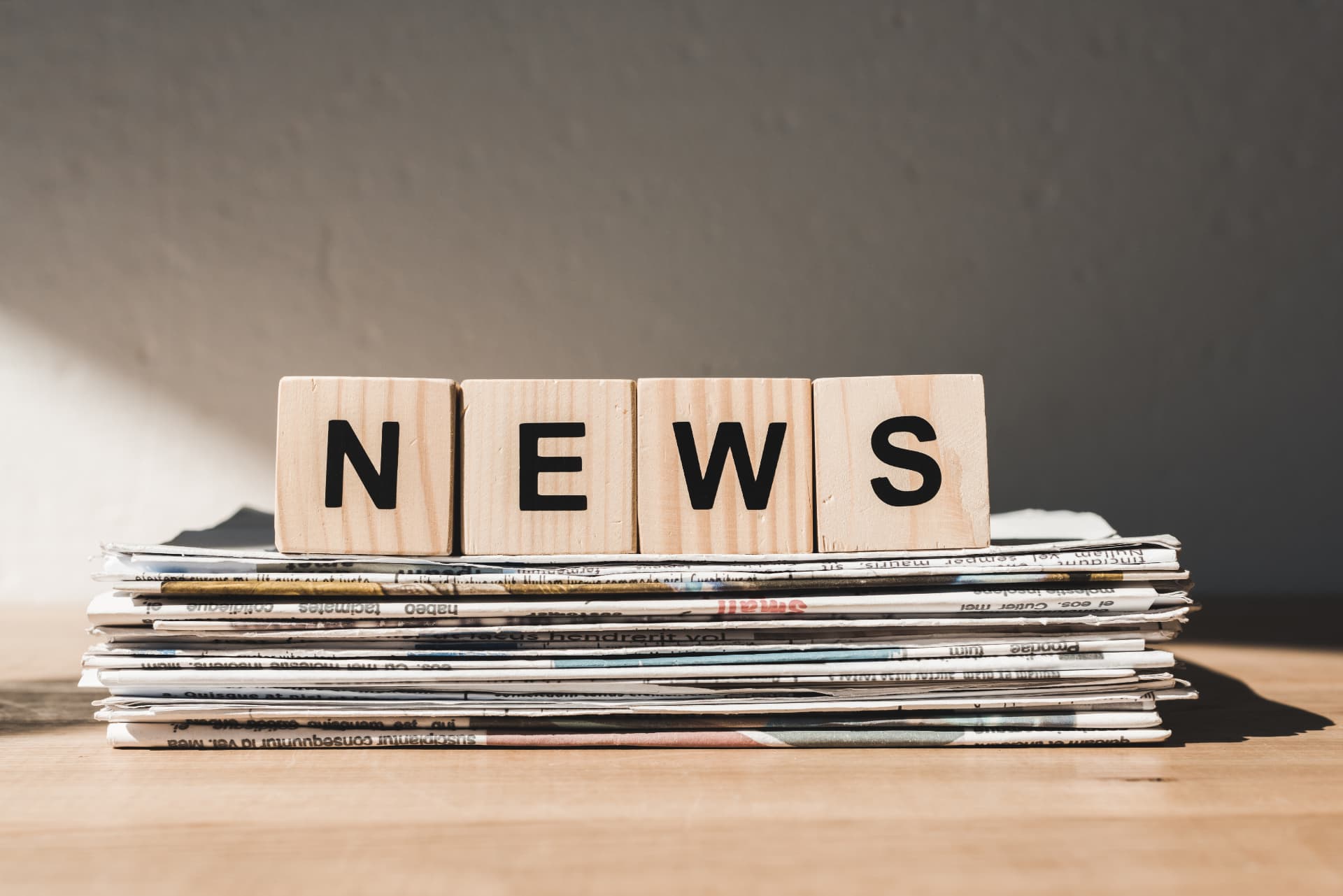 Printed newspapers stacked on a desk beneath wooden letter blocks spelling NEWS — UK Private Wealth Magazine News Desk