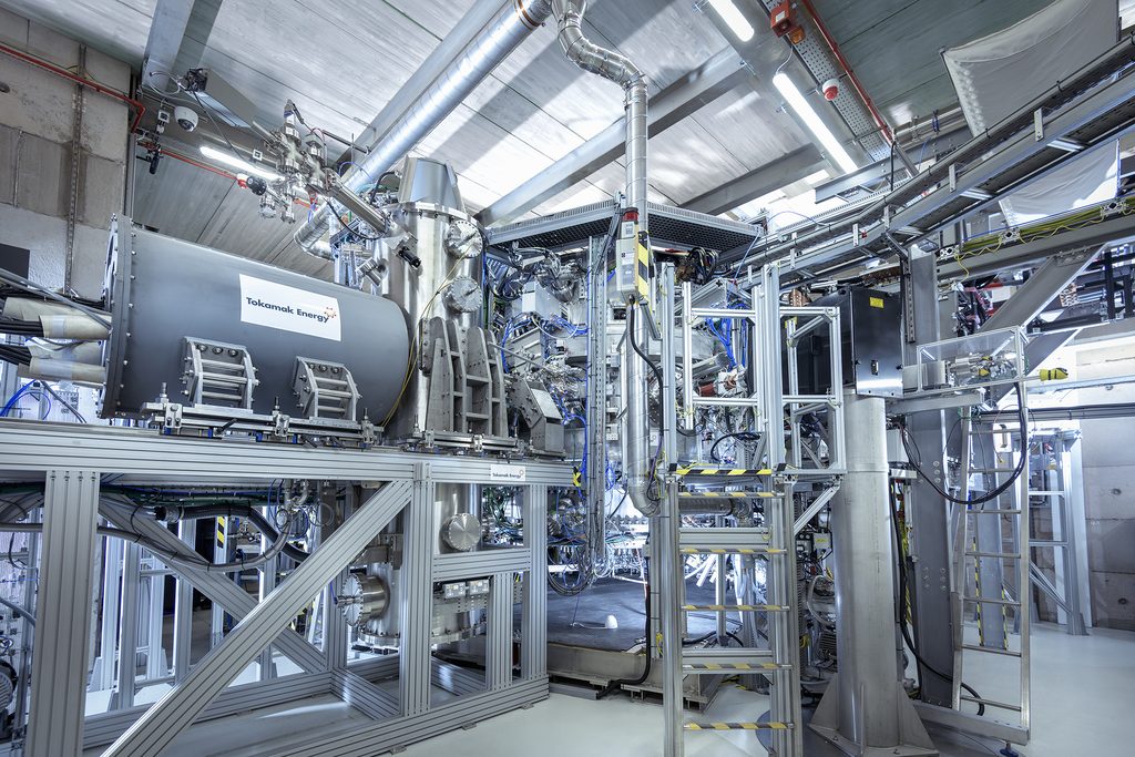 Interior view of Tokamak Energy's ST40 spherical tokamak: a compact steel fusion reactor vessel branded 'Tokamak Energy', surrounded by aluminium support framing, overhead ducting, cabling and access platforms inside the company's experimental hall.