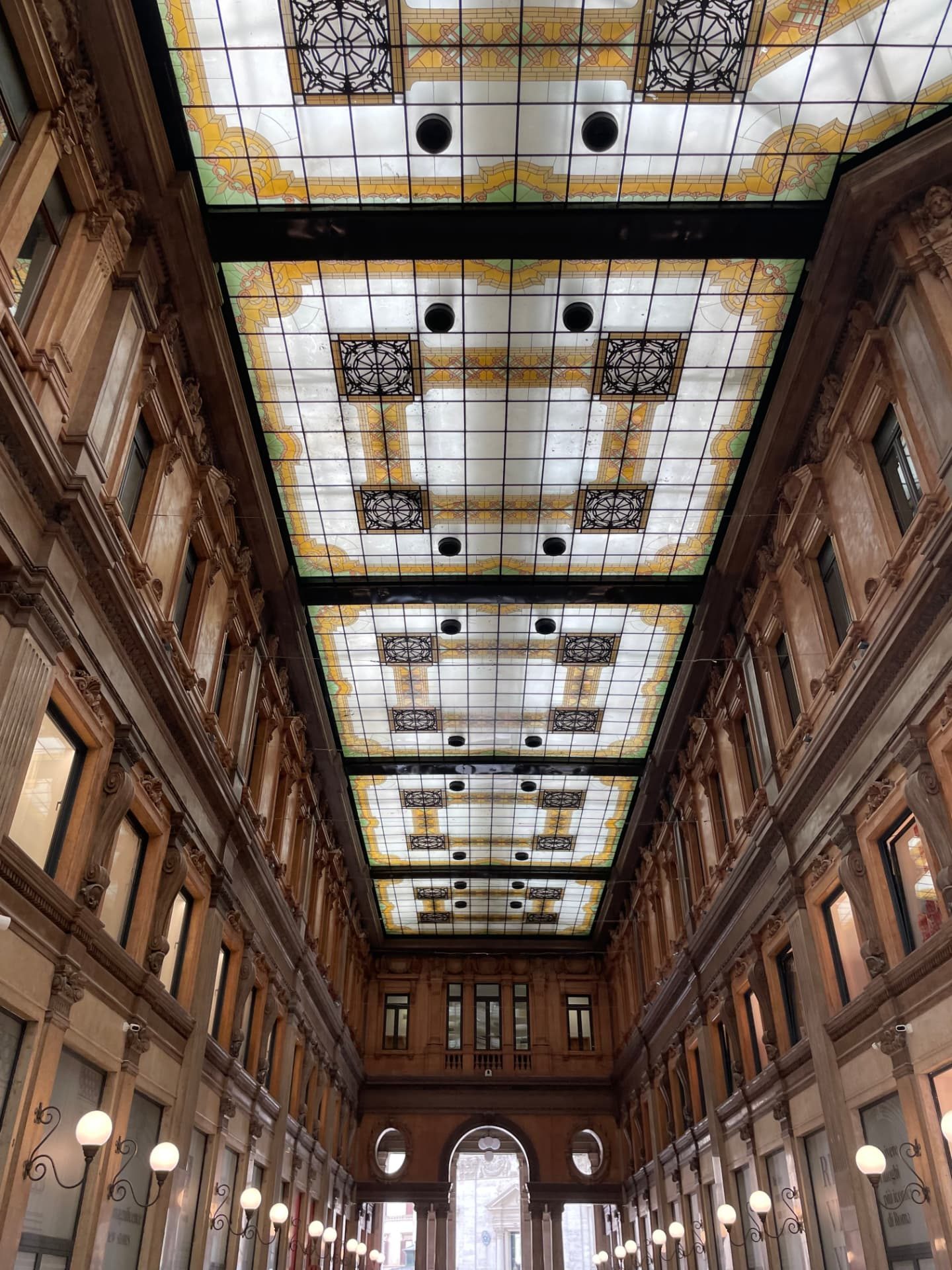 Interior of the Galleria Alberto Sordi in Rome, looking up at the stained-glass barrel skylight above the arcade's stone facades and globe lamps.