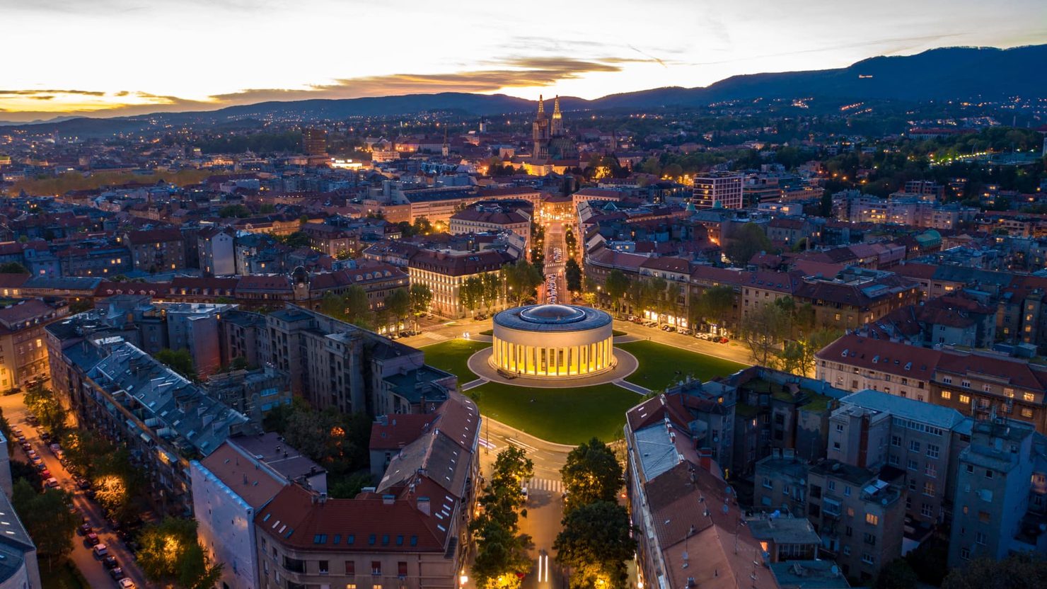 Aerial view of Zagreb, Croatia at dusk, with an illuminated circular building on a broad boulevard among the surrounding city.